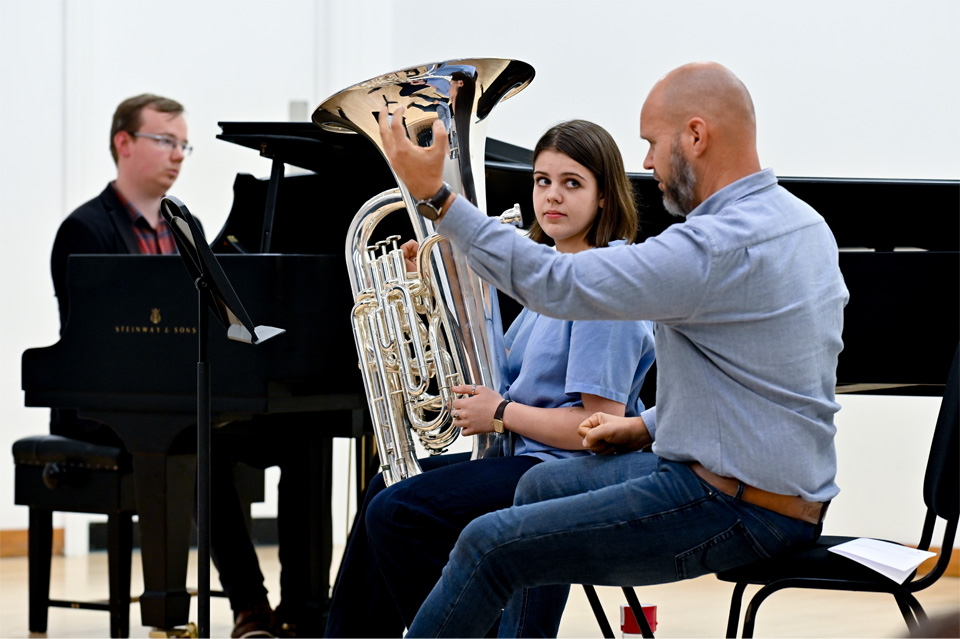 Female student looking at Roland Szentpali, a man in a blue shirt, holding her tuba, while the man teaches her. A piano is in the background to accompany her performance.